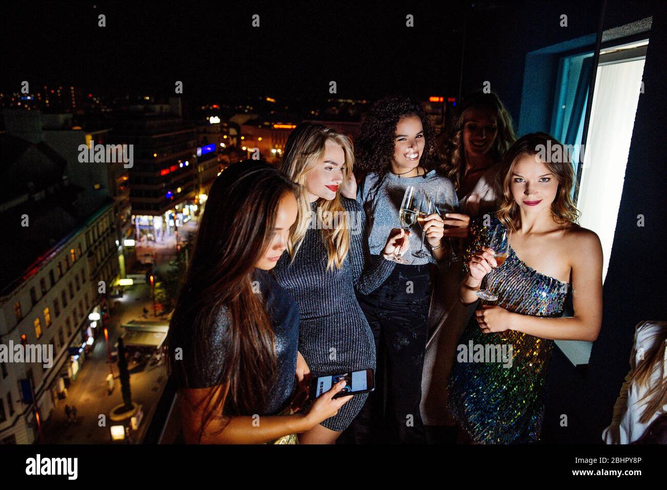 Quatre femmes habillées pour sortir pour la soirée debout dehors sur un balcon avec des verres de champagne. Banque D'Images
