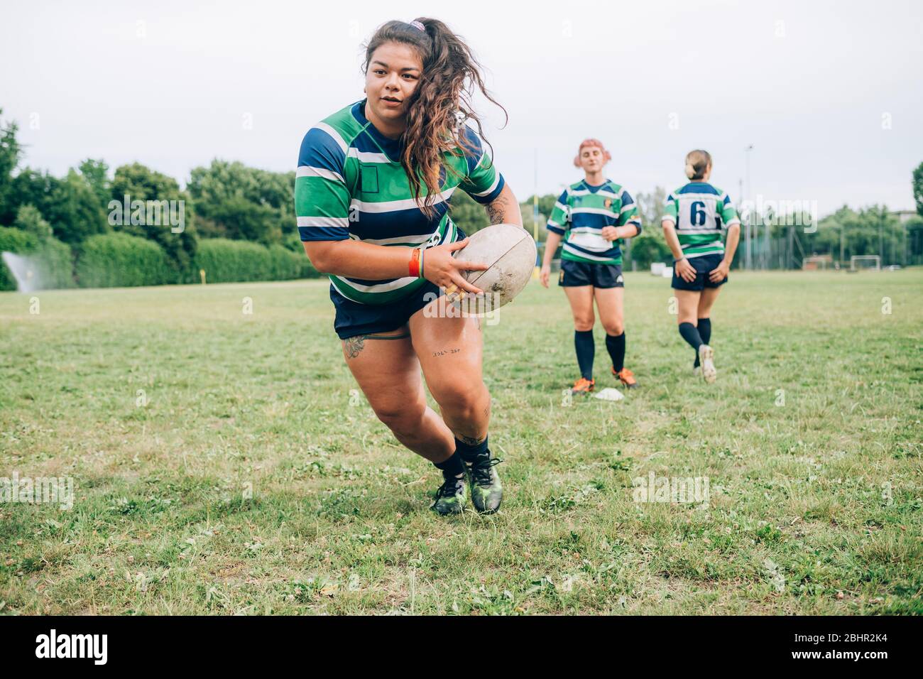 Trois femmes portant des maillots de rugby bleus, blancs et verts sur un terrain d'entraînement, une courir avec un ballon de rugby. Banque D'Images