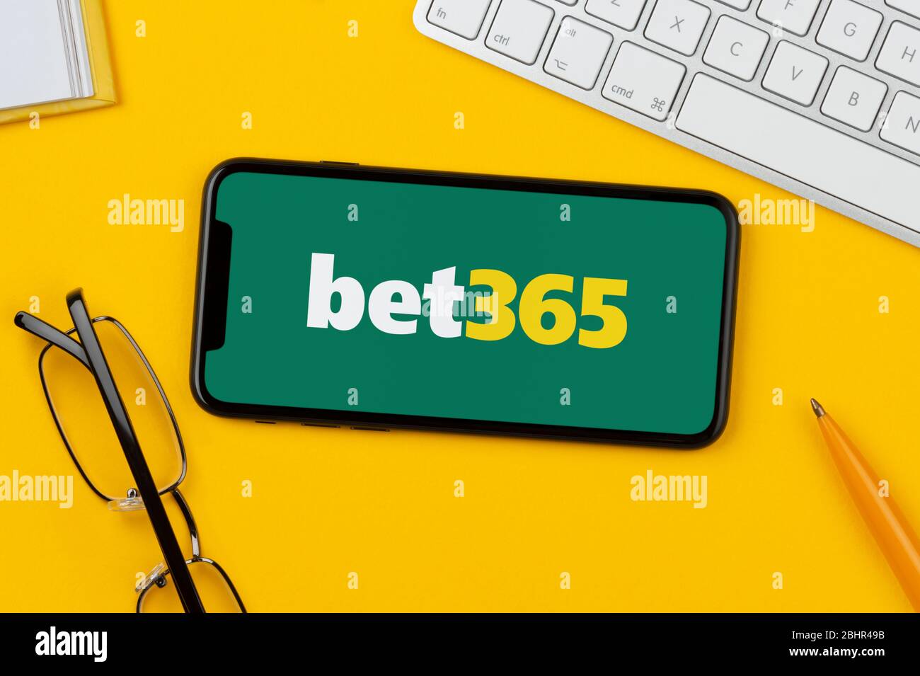 Un smartphone affichant le logo Bet365 repose sur un fond jaune avec un clavier, des lunettes, un stylo et un livre (usage éditorial uniquement). Banque D'Images