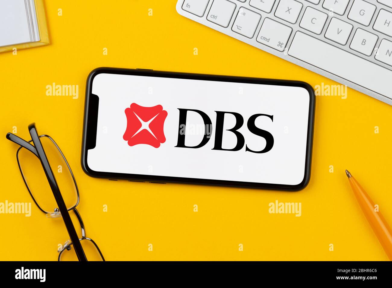 Un smartphone affichant le logo DBS Bank repose sur un fond jaune avec un clavier, des lunettes, un stylo et un livre (usage éditorial uniquement). Banque D'Images