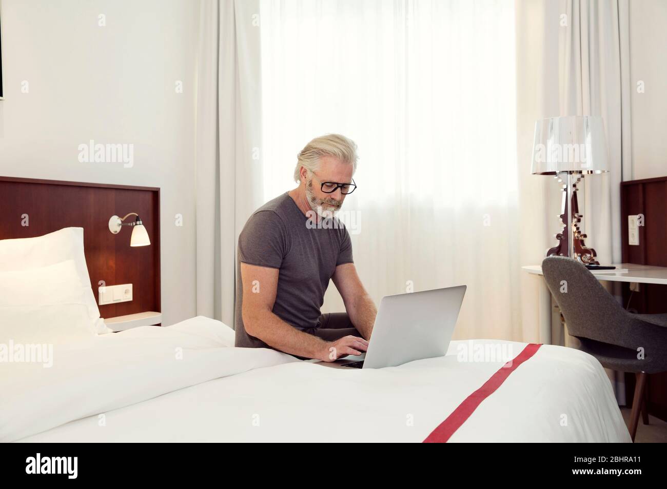 Un homme assis sur un lit qui travaille sur un ordinateur portable. Banque D'Images