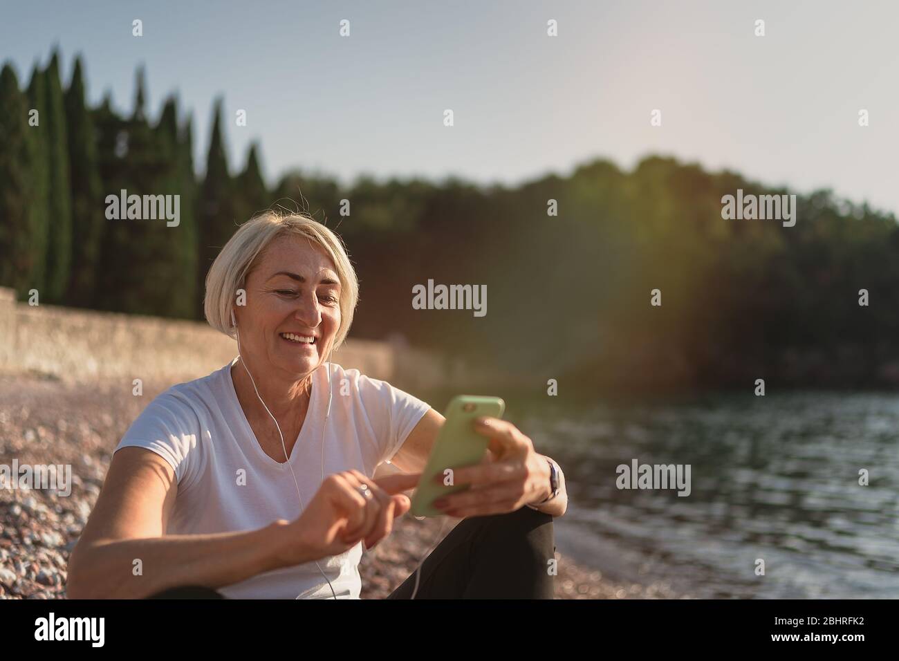Femme mûre assise après le jogging sur la plage. Dame senior utilisant un smartphone et des écouteurs au coucher du soleil. Course en soirée Banque D'Images
