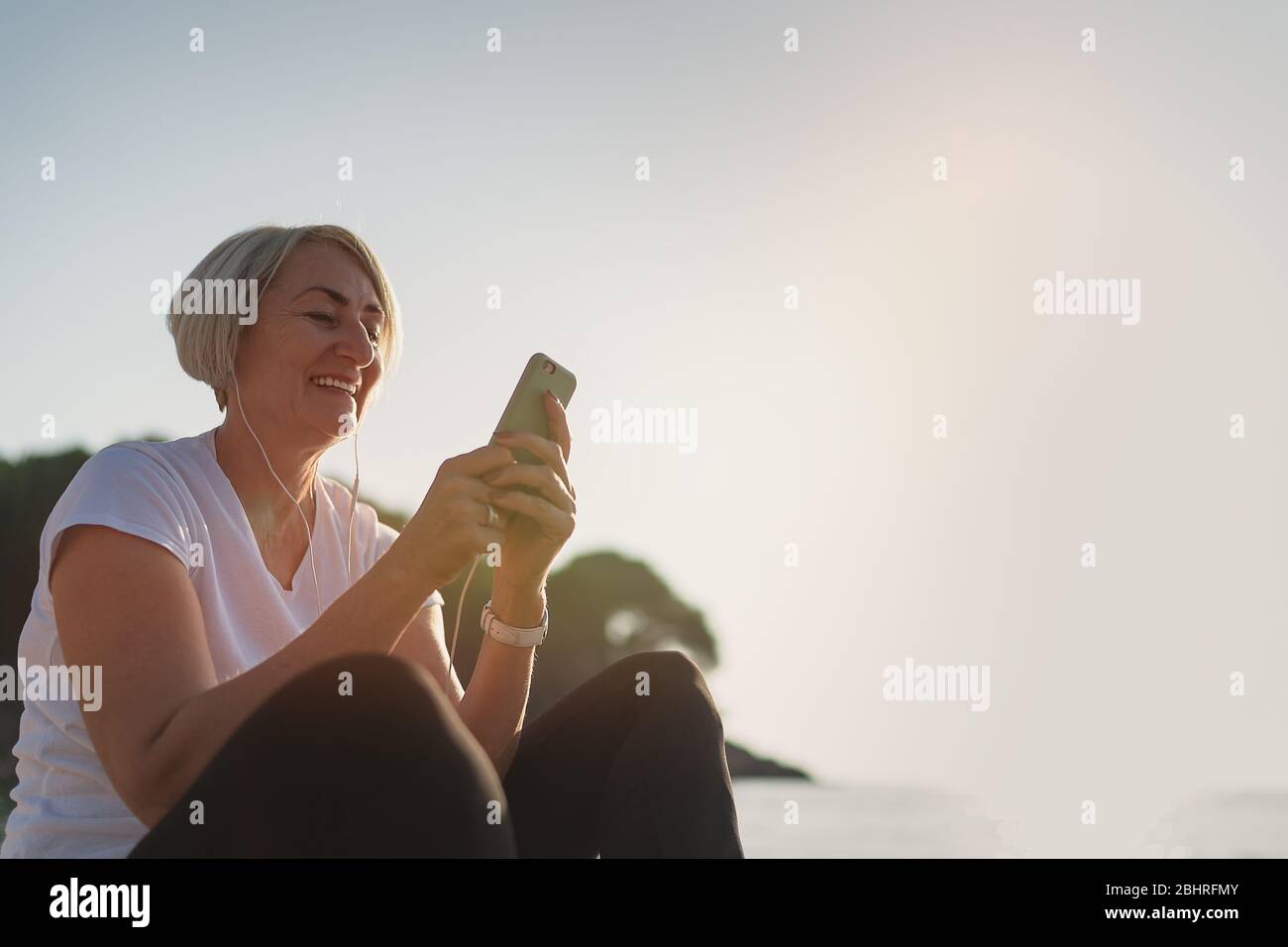 Femme mûre assise après le jogging sur la plage. Dame senior utilisant un smartphone et des écouteurs au coucher du soleil. Course en soirée Banque D'Images