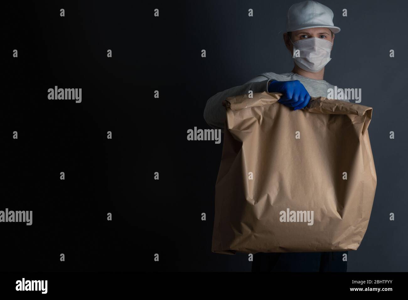 Livraison sûre de nourriture ou de marchandises. Un jeune transporteur livre à la maison du client une grosse commande de sacs en papier vert brun avec masque et gants pendant la co Banque D'Images