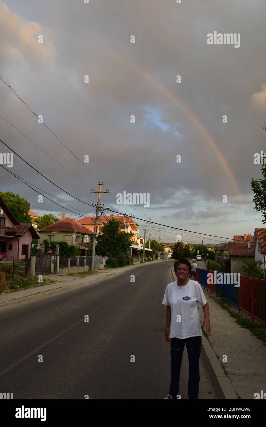 Une femme âgée se tient sur le pavé à côté du trottoir. En arrière-plan sont des maisons de campagne et un ciel gris avec un arc-en-ciel Banque D'Images