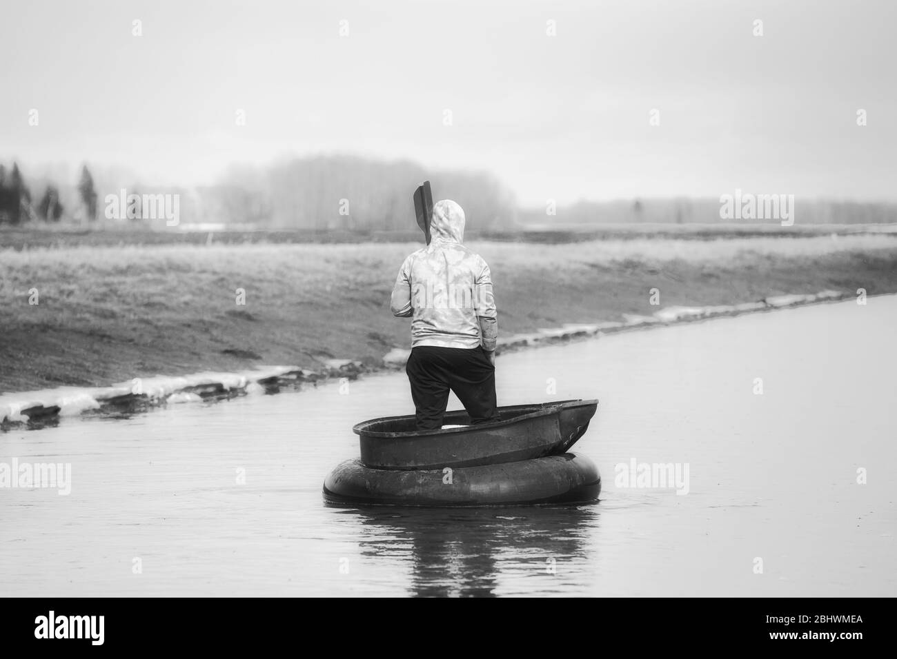 L'arrière d'un jeune homme debout dans un tube de pneu avec une palette sur son épaule flottant vers le bas d'un fossé dans un paysage de printemps noir et blanc Banque D'Images