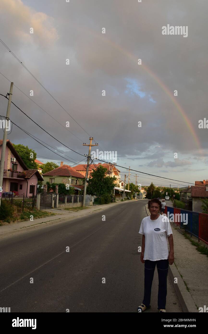 Une femme âgée se tient sur la route près du trottoir. Derrière la femme, il y a un arc-en-ciel dans le ciel Banque D'Images