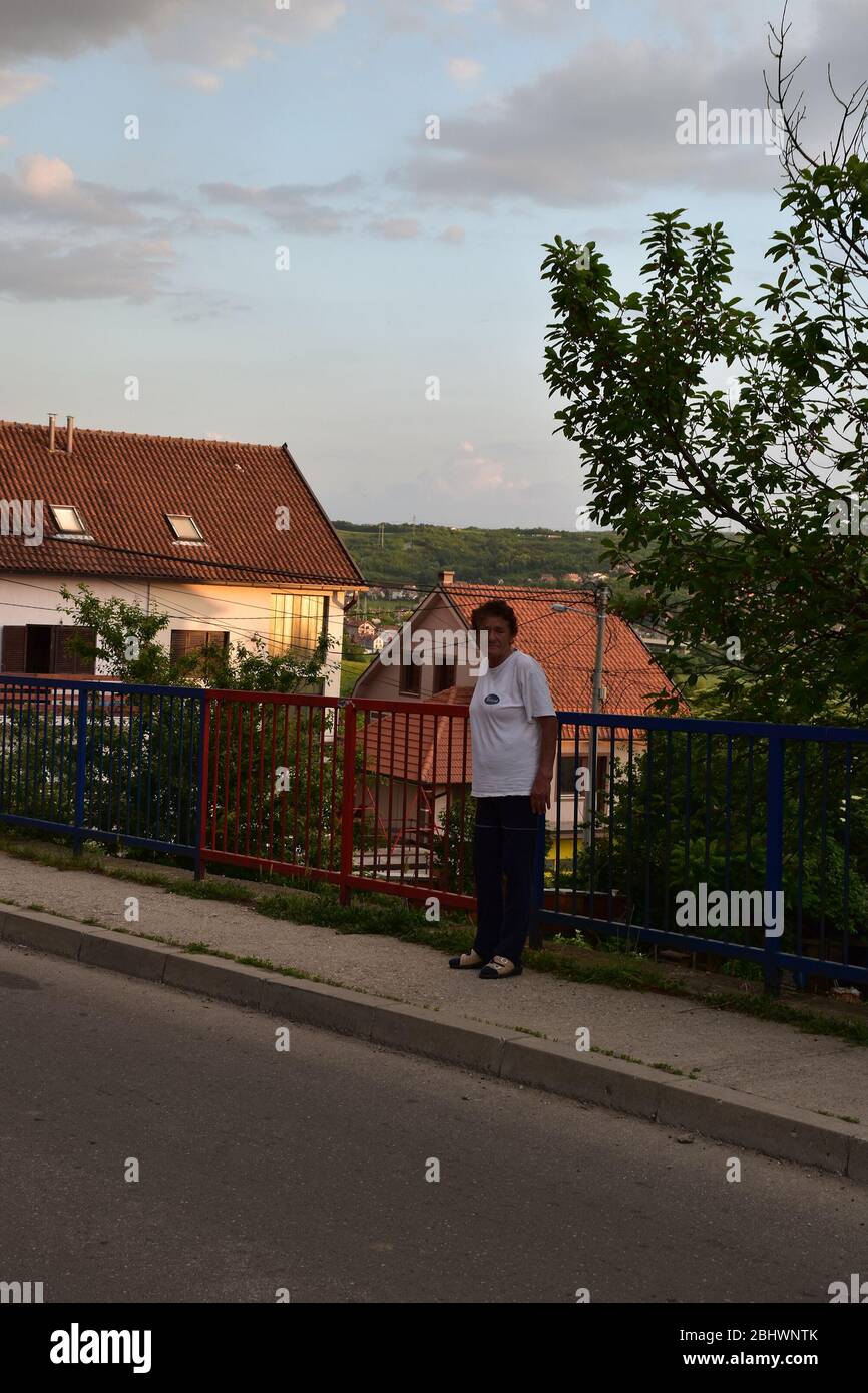 Sur le trottoir, sur le côté de la route, une femme plus âgée se tient. En arrière-plan sont les maisons et la nature Banque D'Images