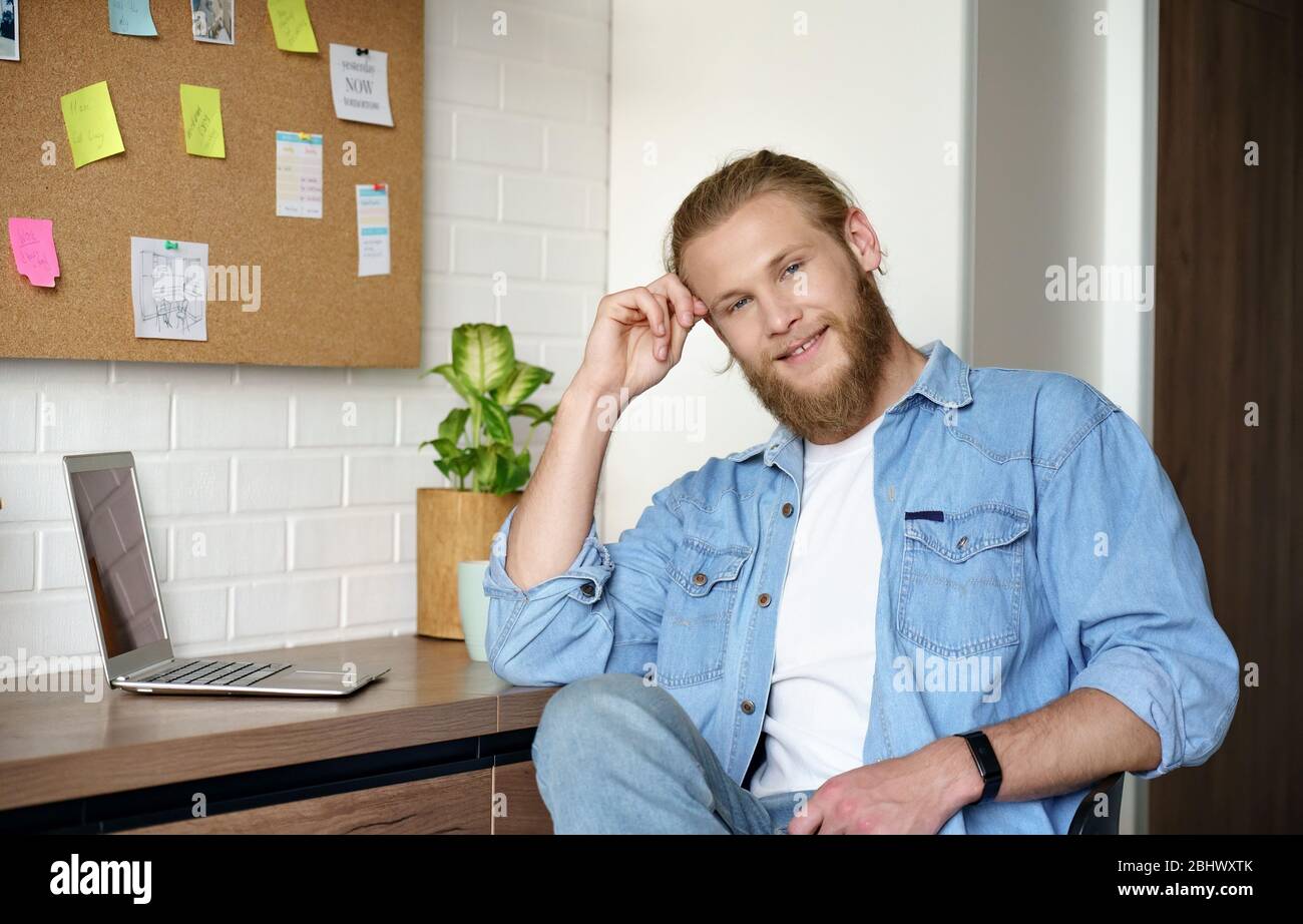 Jeune homme souriant regardant un appareil photo assis à la maison bureau avec ordinateur portable. Banque D'Images