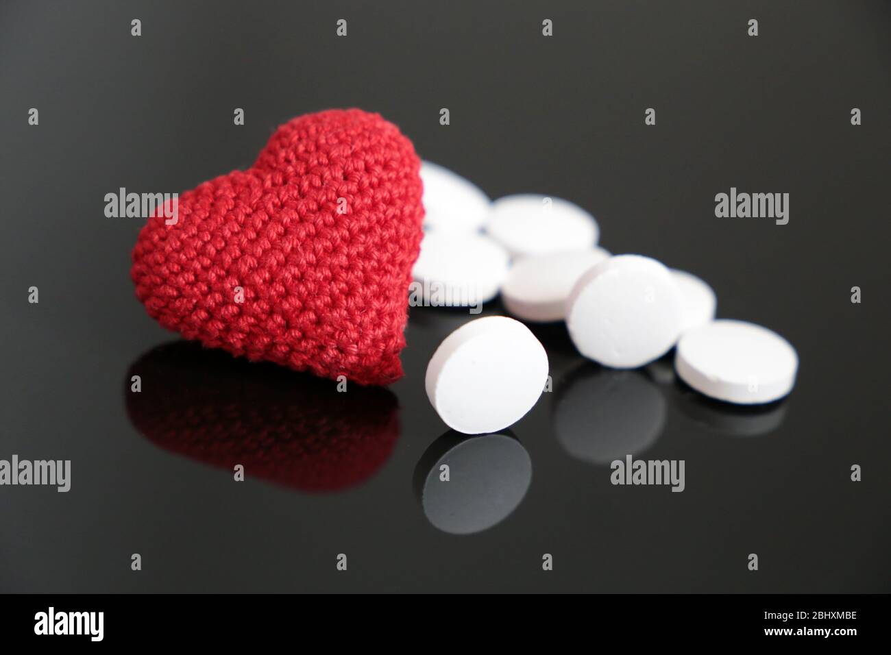 Pilules blanches et coeur rouge tricoté sur table en verre sombre. Concept d'hypertension, de maladie cardiaque, de cardiologie Banque D'Images