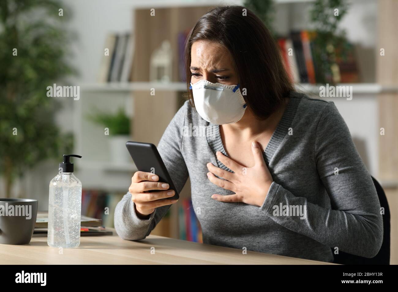 Une femme effrayée avec masque de protection due coronavirus lit des nouvelles en ligne sur un smartphone assis sur un bureau dans la nuit à la maison Banque D'Images