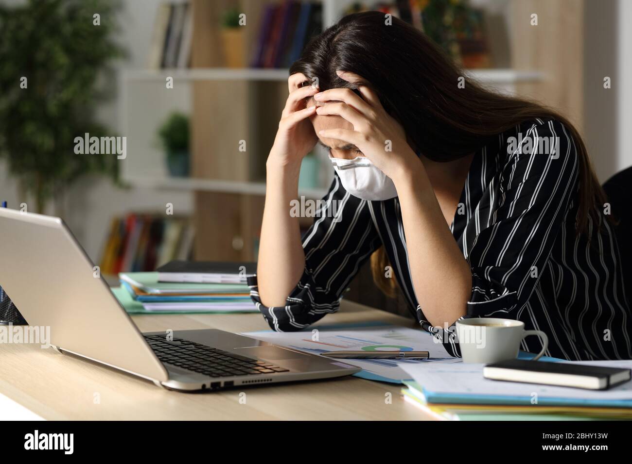 Triste télétravailleur femme portant un masque avec des problèmes dus coronavirus assis sur un bureau avec ordinateur portable dans la nuit au bureau à la maison Banque D'Images