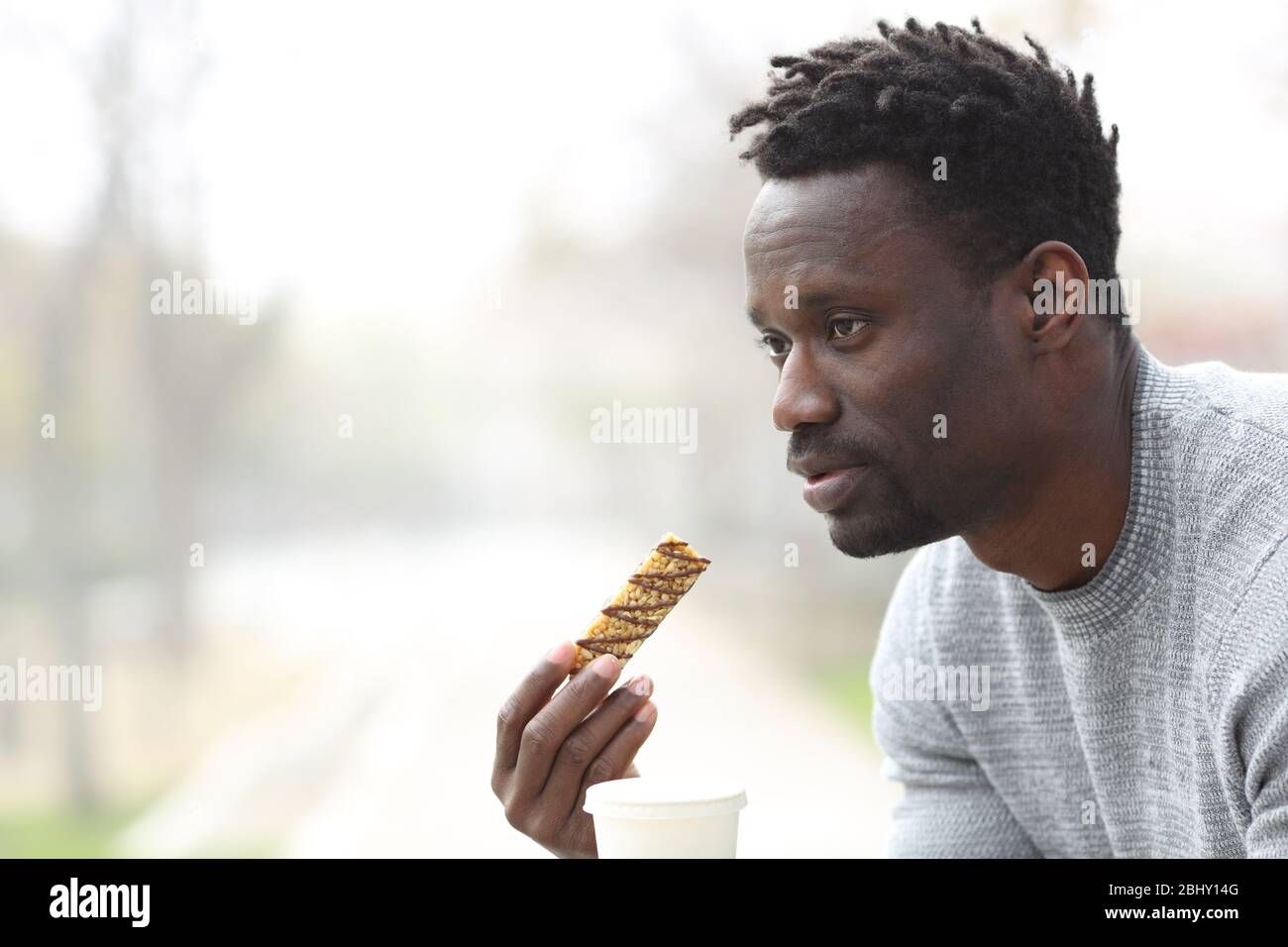 Homme noir sérieux avec snack-bar à céréales et café à emporter assis sur un banc de parc Banque D'Images