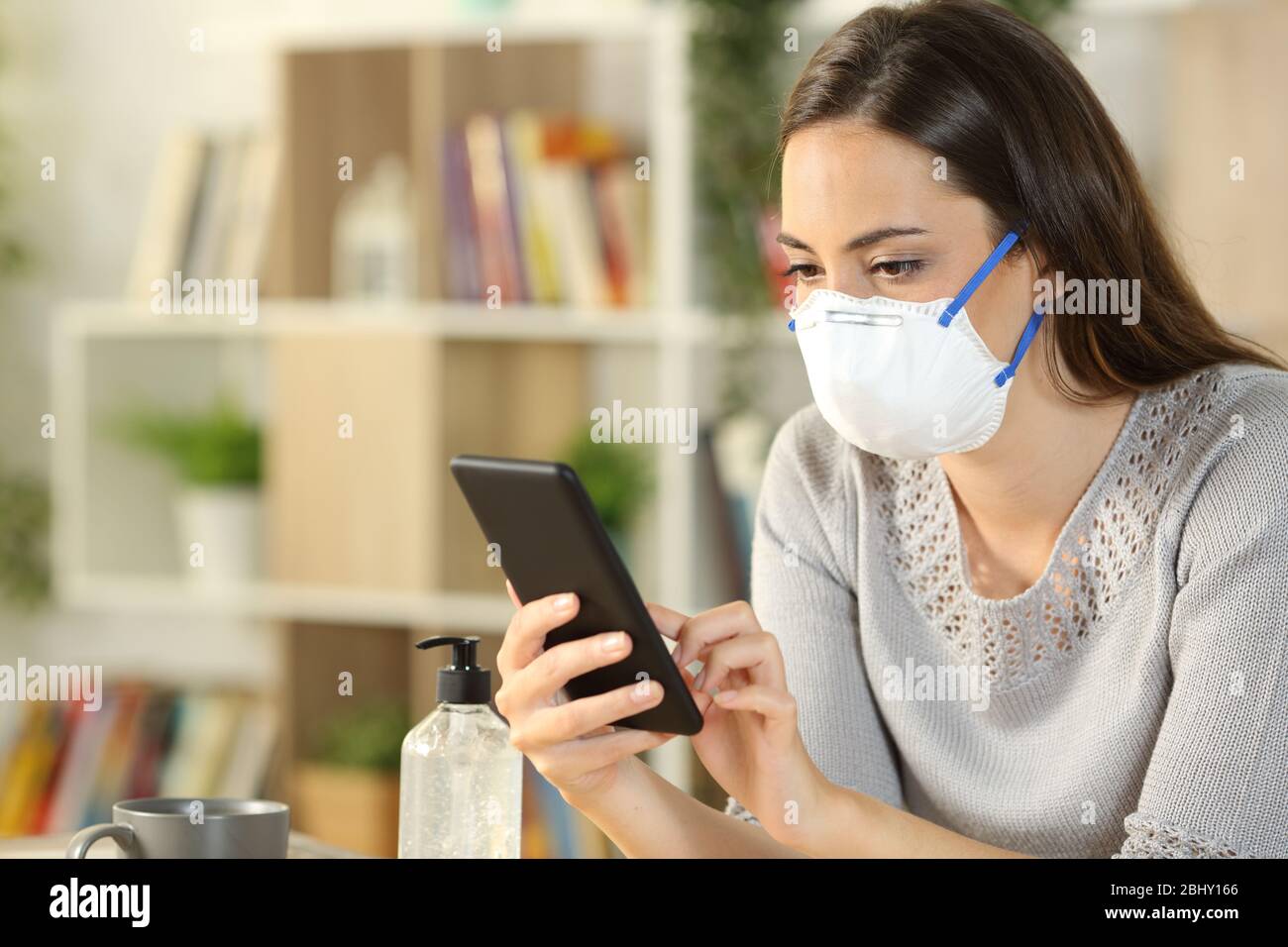 Femme sérieuse avec masque de protection dû coronavirus utilisant un smartphone assis sur le salon à la maison Banque D'Images