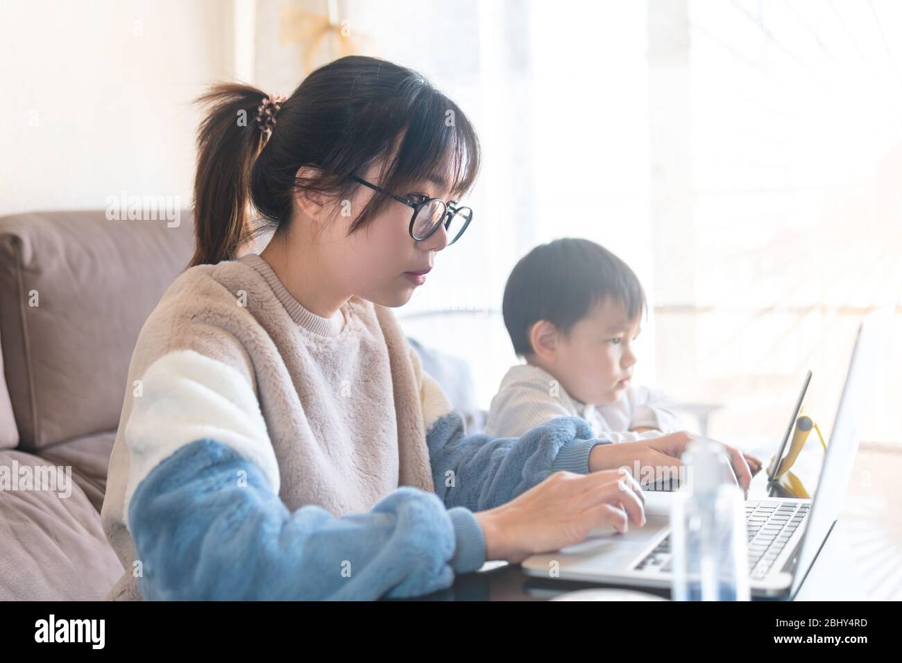 Jeune mère asiatique travaillant à domicile sur ordinateur. Un enfant regarde un dessin animé sur une tablette tandis que sa mère travaille sur un ordinateur portable pendant le blocage de la pandémie du coronavirus Banque D'Images
