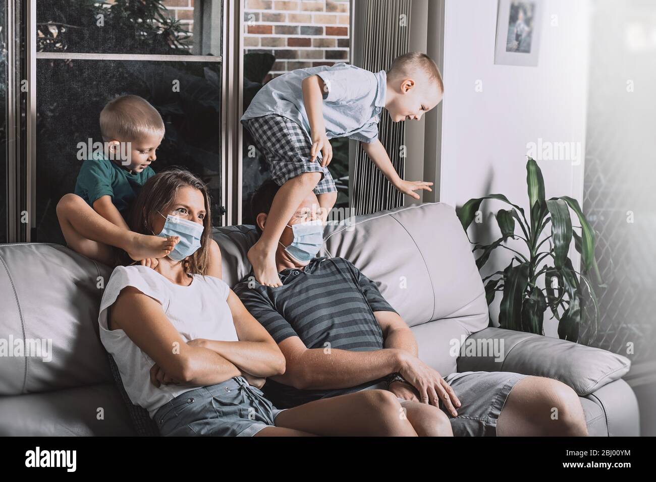 Maman et papa portent des masques chirurgicaux pour le visage. Deux petits fils sautaient sur les parents assis sur le canapé pour essayer de regarder la télévision. Toute famille reste à la maison pendant le verrouillage de la quarantaine. Banque D'Images