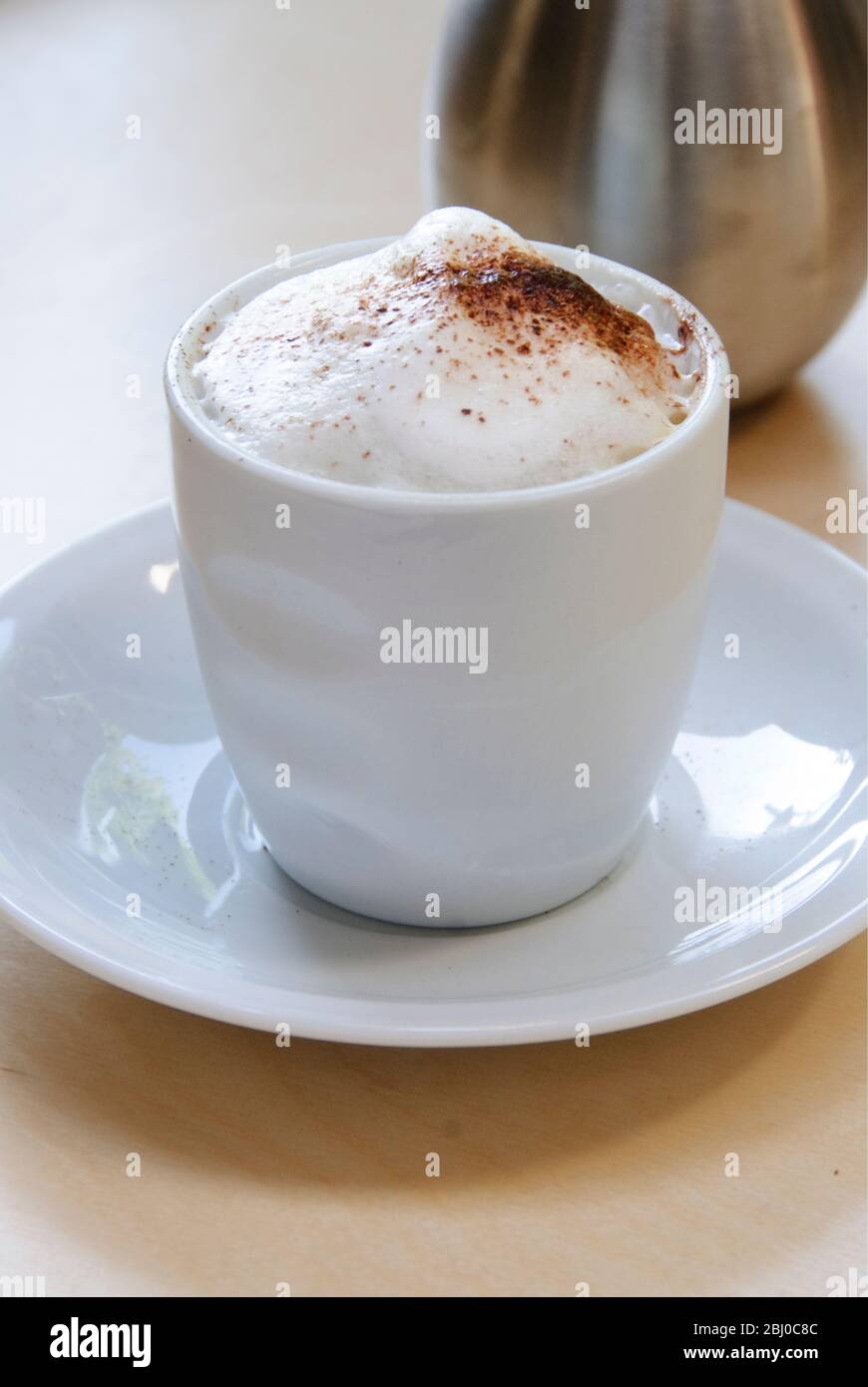 Cappuccino surmonté de mousse dans une tasse sans main avec du cacao dépoli et un shaker de cacao derrière - Banque D'Images