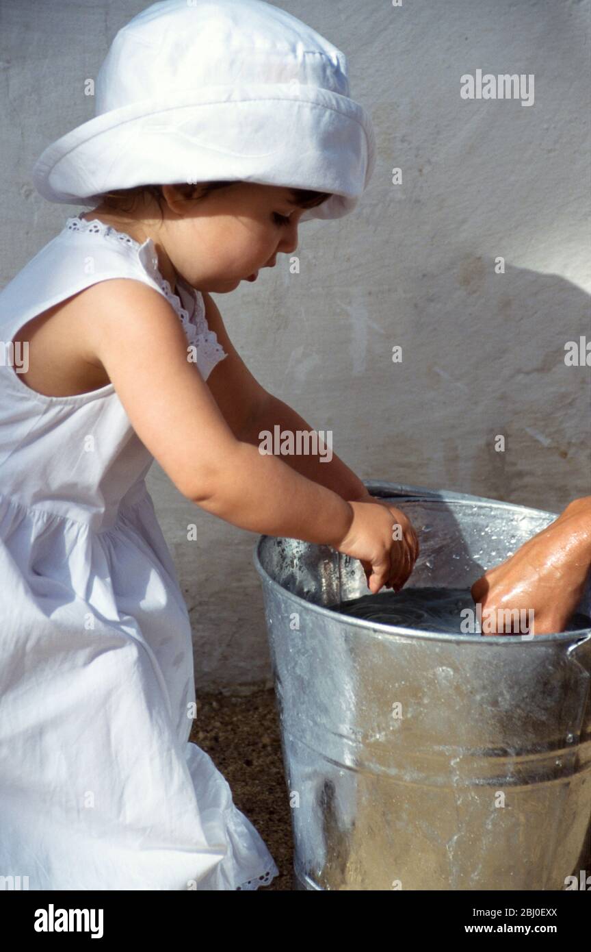 Petite fille jouant avec un seau d'eau claire et quelques chevilles de vêtements alors que la mère est pendante du lavage dans la cour ensoleillée - Banque D'Images