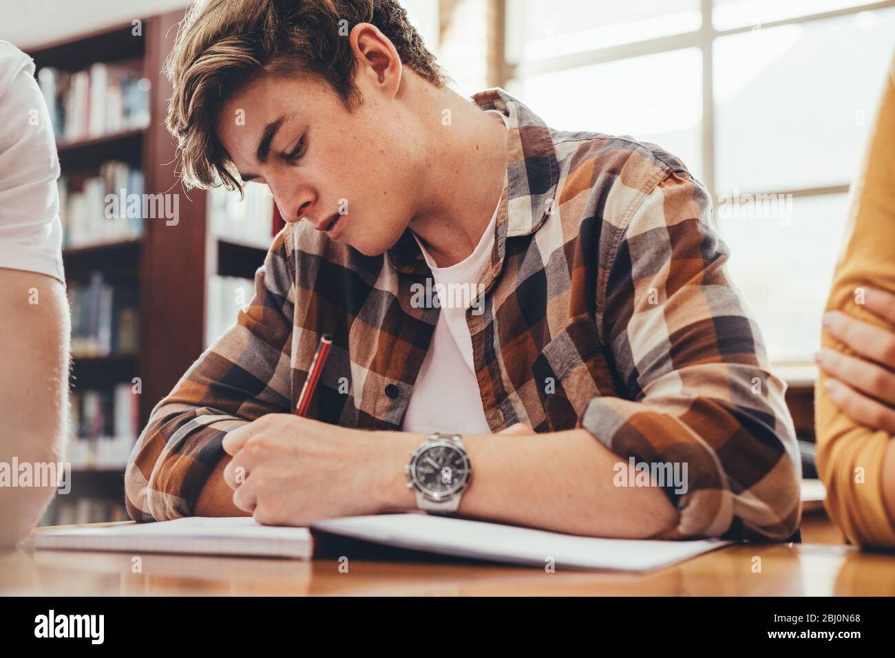 Étudiant masculin assis à la table de bibliothèque et écrivant des notes dans un livre. Étudiant étudiant étudiant dans une bibliothèque de lycée avec des camarades de classe assis autour. Banque D'Images