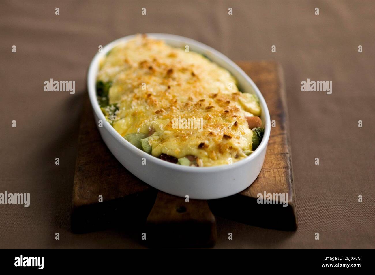 Poireaux et jambon mornay, poireaux enveloppés dans du jambon avec une sauce au fromage dans un plat blanc, ovale et ovenimeux sur planche en bois - Banque D'Images