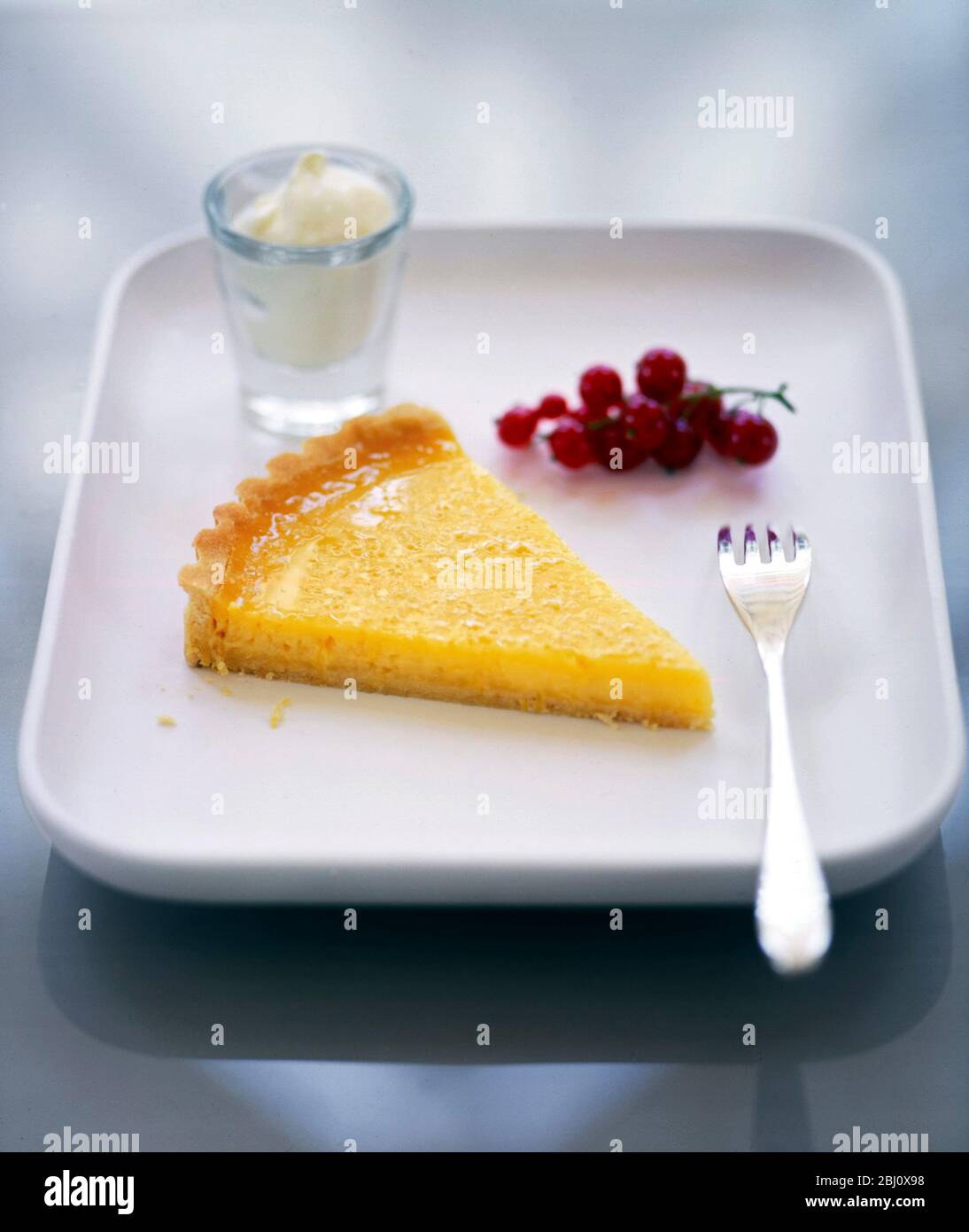 Tranche de tarte au citron classique servie avec des groseilles fraîches, du zeste de citron et de la crème fraîche effilochée - Banque D'Images