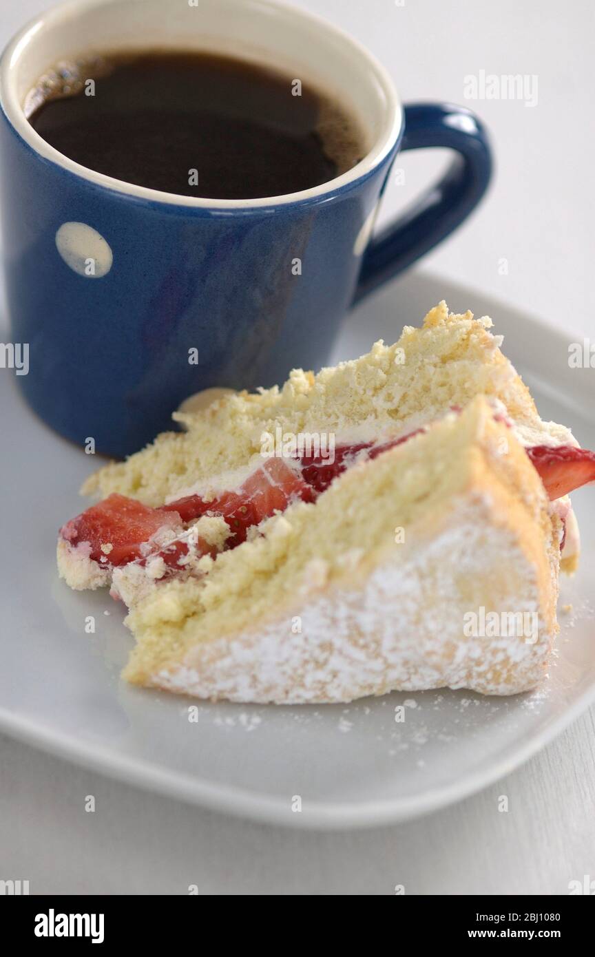 Un morceau de gâteau éponge de fraise sur une plaque blanche avec un petit mug à pois bleu et blanc de café noir - Banque D'Images