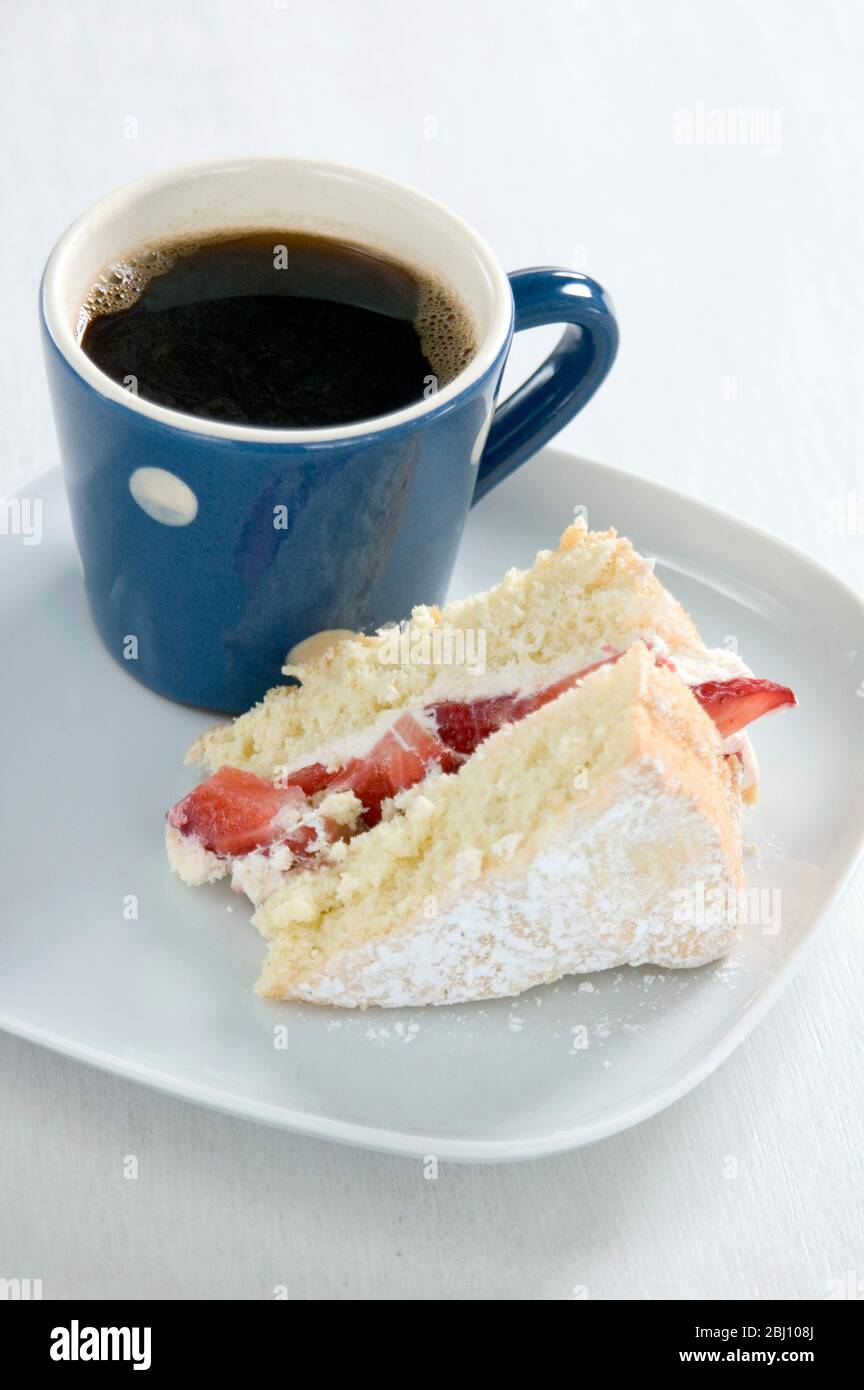 Un morceau de gâteau éponge de fraise sur une plaque blanche avec un petit mug à pois bleu et blanc de café noir - Banque D'Images
