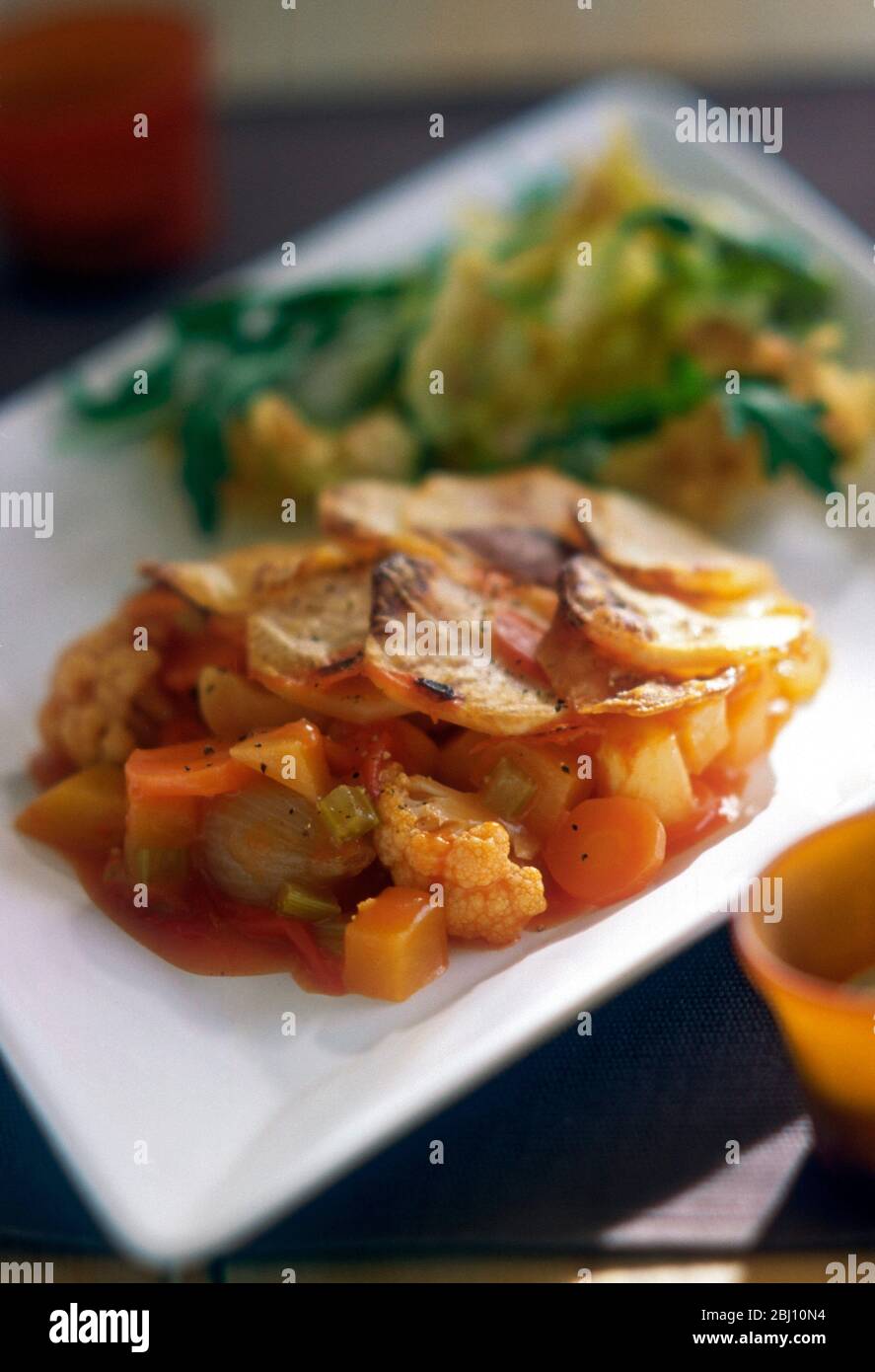 Plat végétarien chaud composé de légumes mélangés avec garniture de pommes de terre en tranches, servi avec salade de roquette sur une plaque blanche carrée moderne. - Banque D'Images