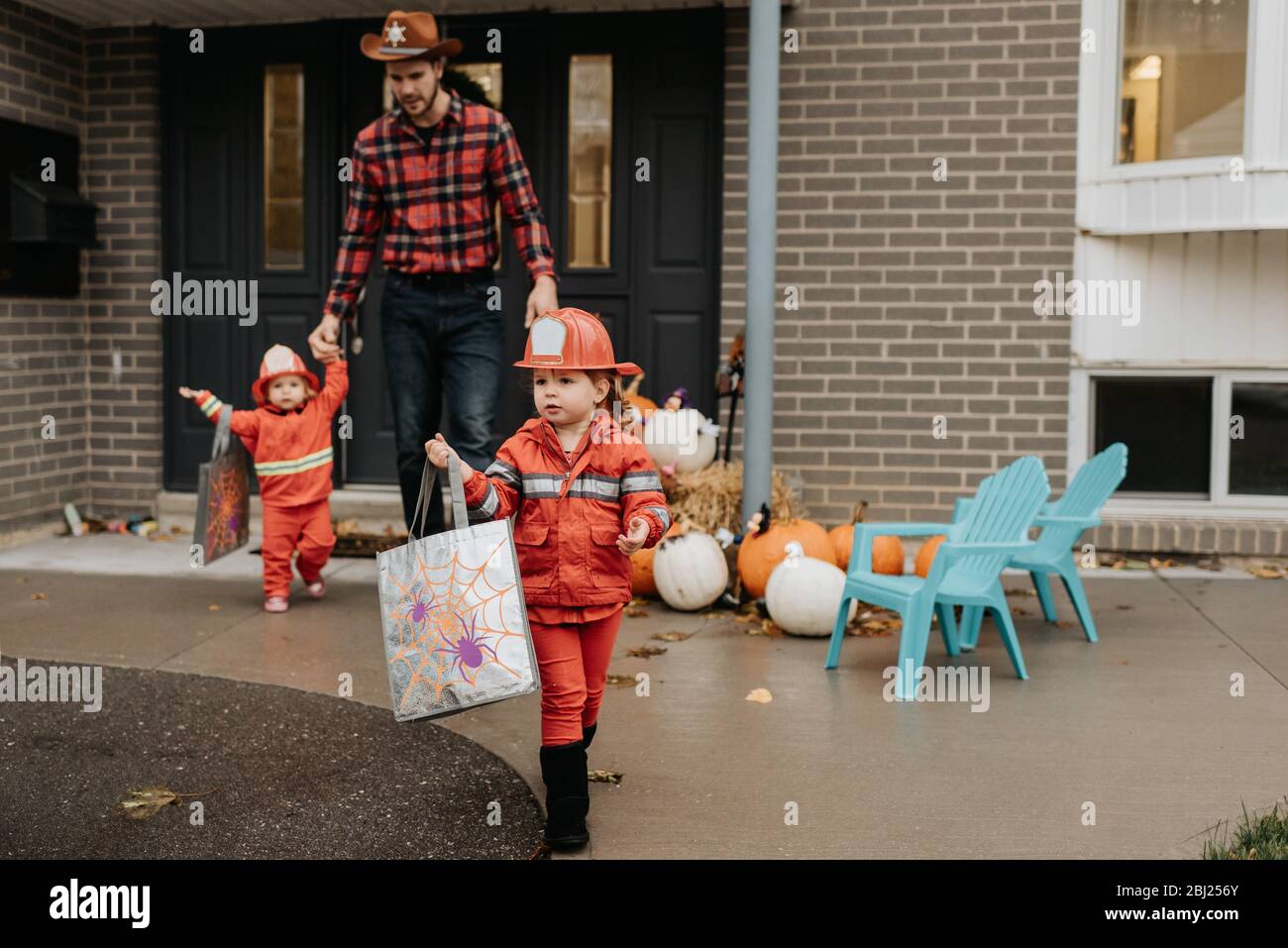 Deux enfants vêtus de pompiers marchant loin de la porte d'entrée d'une maison décorée pour Halloween avec leur père. Banque D'Images