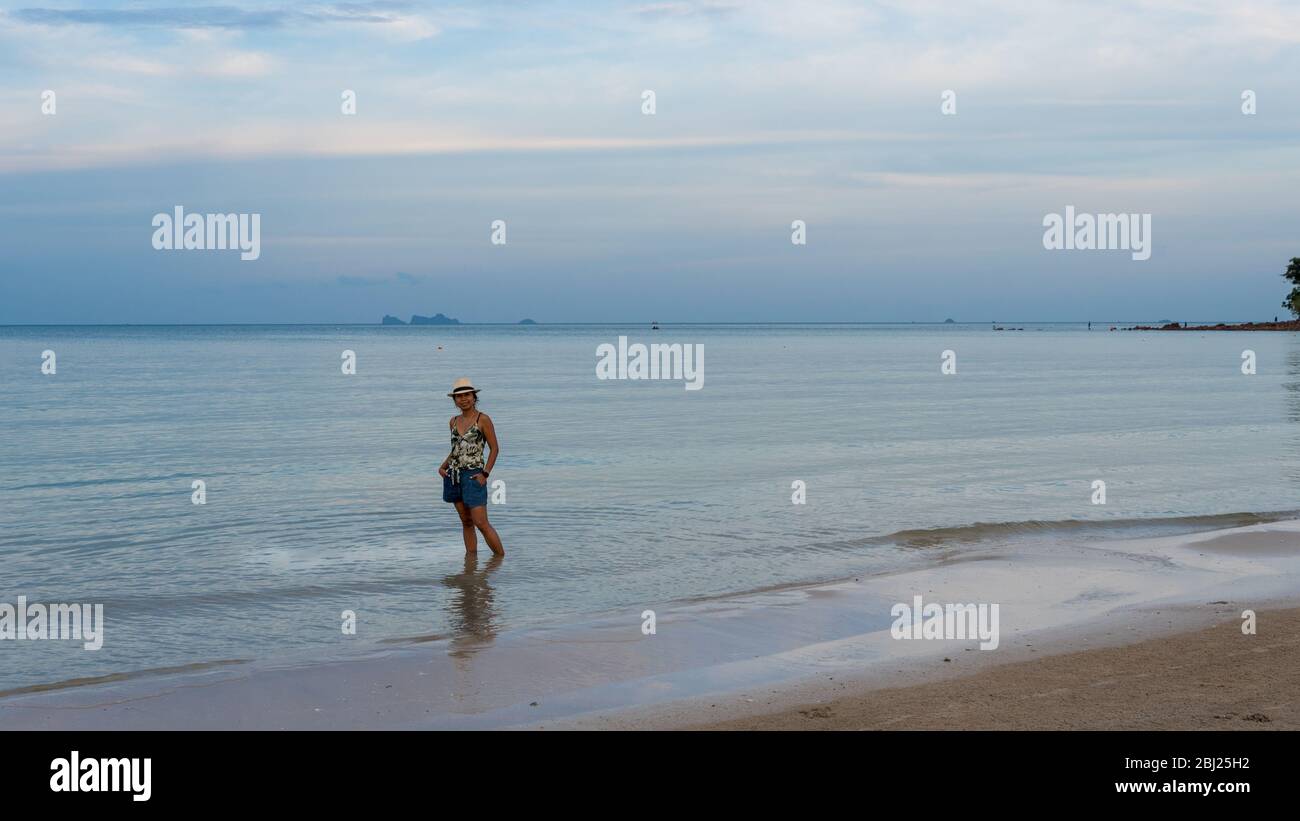 Femme thaïlandaise en short et chapeau debout dans l'eau peu profonde pieds nus sur la plage à marée basse à Pythiu sur le golfe de Thaïlande Banque D'Images
