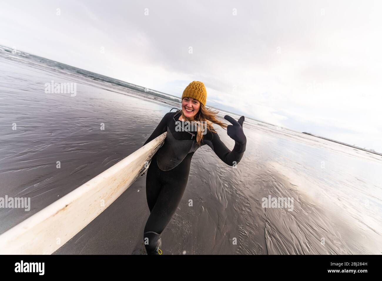 Une femme portant une combinaison et portant une planche de surf marchant  sur une plage noire avec la mer en arrière-plan Photo Stock - Alamy