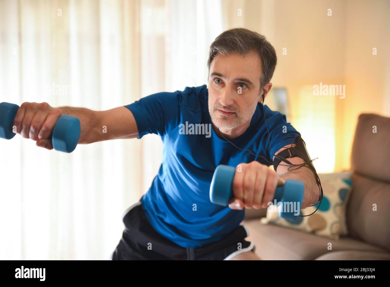 Homme faisant du sport avec des haltères dans le salon à la maison écoutant de la musique avec des écouteurs et le téléphone Banque D'Images