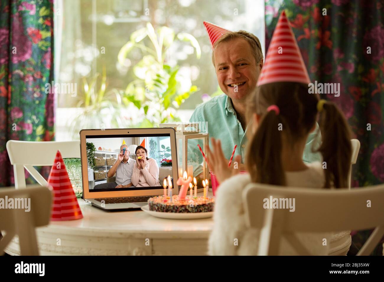 Bonne petite fille célébrant son anniversaire à la maison avec ses parents et ses grands parents par vidéoconférence. Ordinateur portable avec couple senior en ligne, gâteau avec bougies Banque D'Images