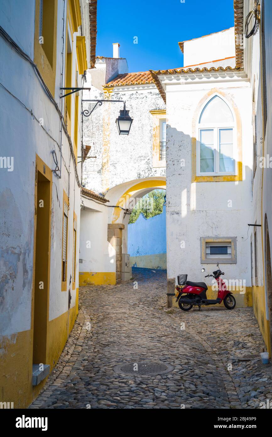 Scooter à moteur stationné dans une rue calme en pierre de galets par des maisons typiques blanches et jaunes à Evora, Portugal Banque D'Images