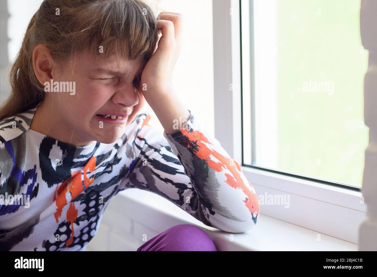 Une jeune fille pleurant beaucoup en s'assissant près de la fenêtre dans la chambre, gros plan Banque D'Images