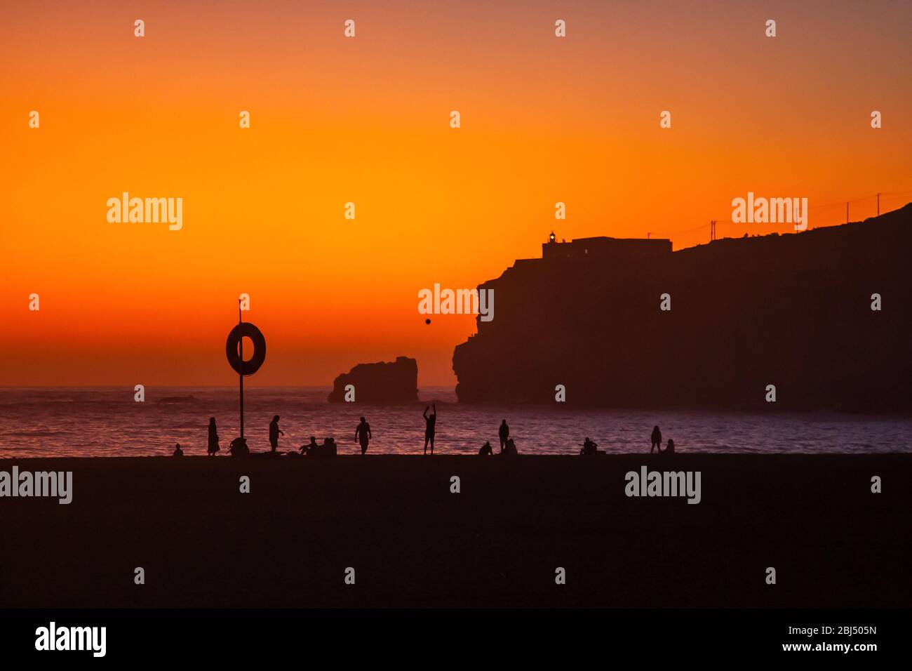 Nazaré, Portugal : magnifique coucher de soleil orange sur l'océan Atlantique et les gens se détendent sur la plage. Phare et fort de Sao Miguel visibles sur la haute Banque D'Images