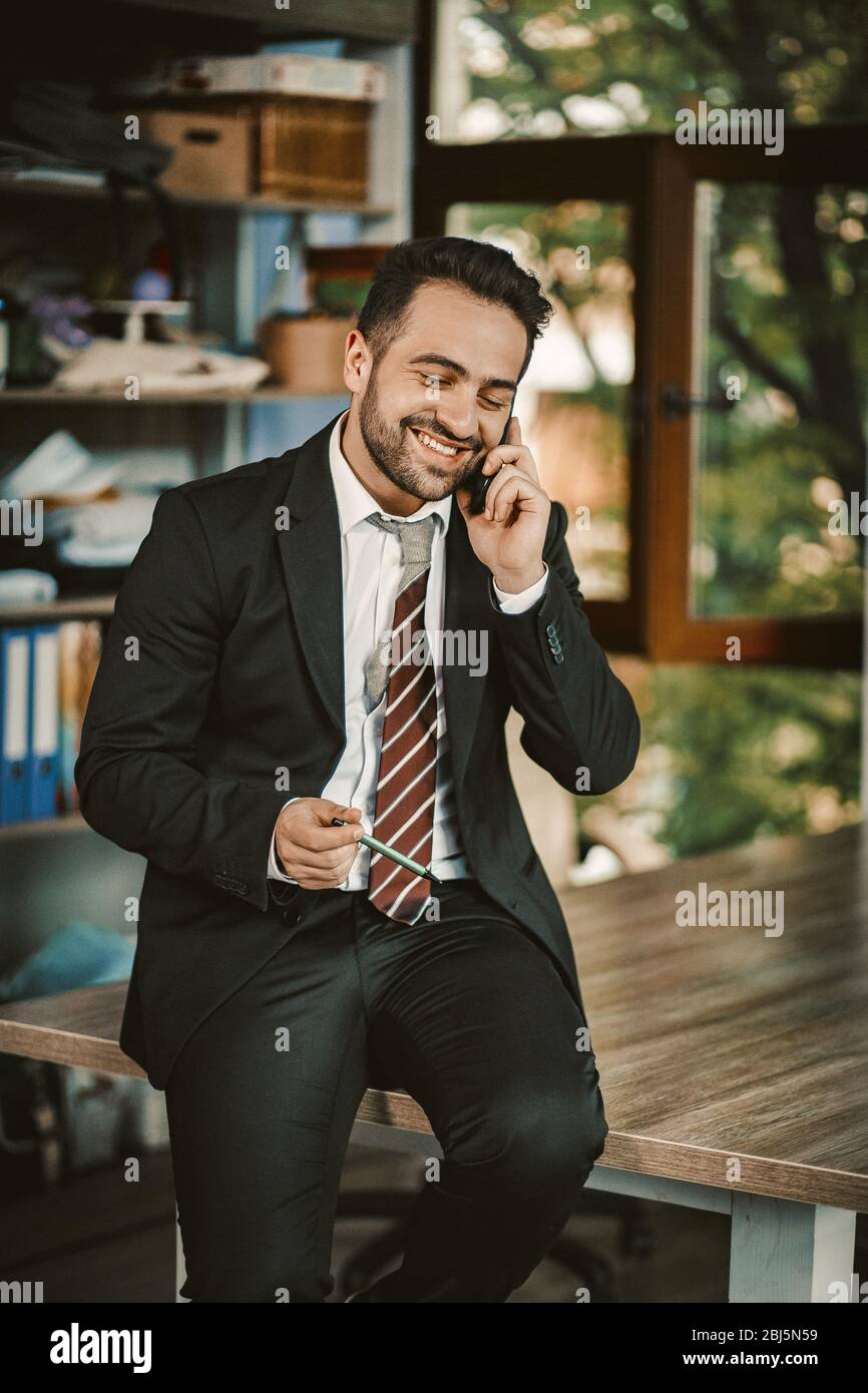 Un homme d'affaires joyeux et joyeux parle avec le téléphone portable assis sur le bureau Banque D'Images