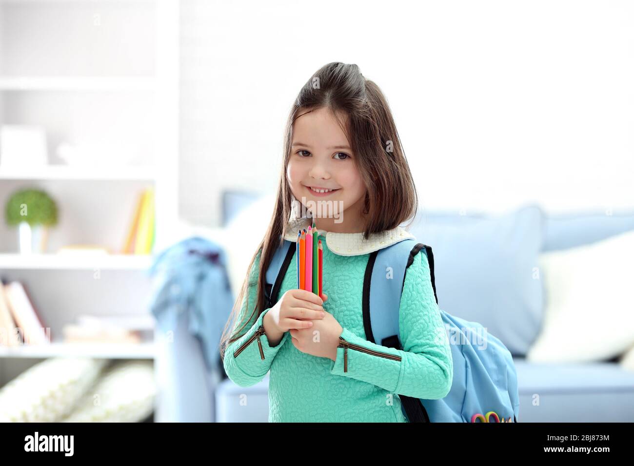 Petite fille avec sac à dos bleu tenant des crayons de couleur dans le salon Banque D'Images