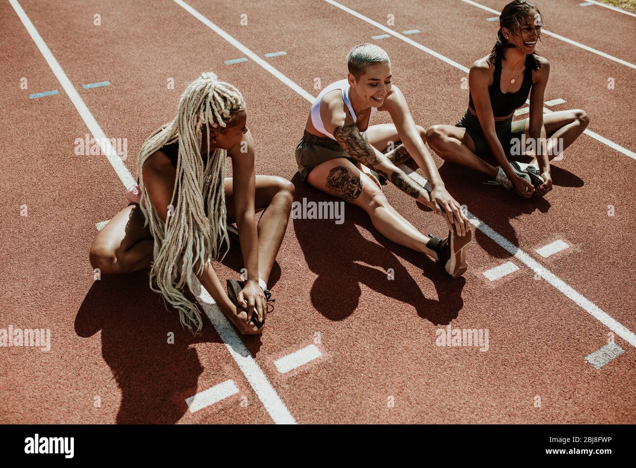 Trois athlètes féminins assis sur une piste de course et des jambes étirements. Groupe multiethnique de coureurs faisant des exercices de préchauffage au stade. Banque D'Images