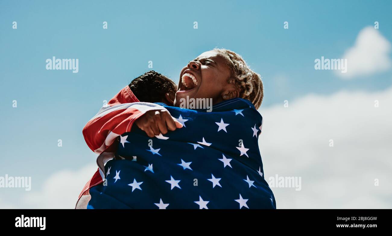 Les athlètes féminins américains excités enveloppés dans un drapeau national s'embrassant après une victoire. L'équipe des coureurs de femmes des États-Unis se réjouit d'une victoire Banque D'Images