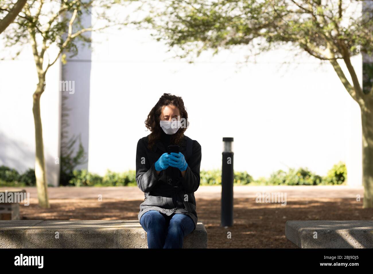 Femme caucasienne portant un masque de protection et des gants assis et utilisant son téléphone Banque D'Images