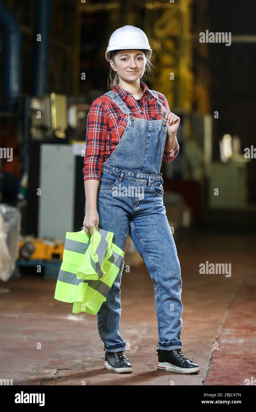 Jeune fille dans une robe de travail et un chapeau blanc dans une usine.  Femme dans un uniforme de travail tenant un gilet réfléchissant dans un  rangement. Processus de travail Photo Stock -