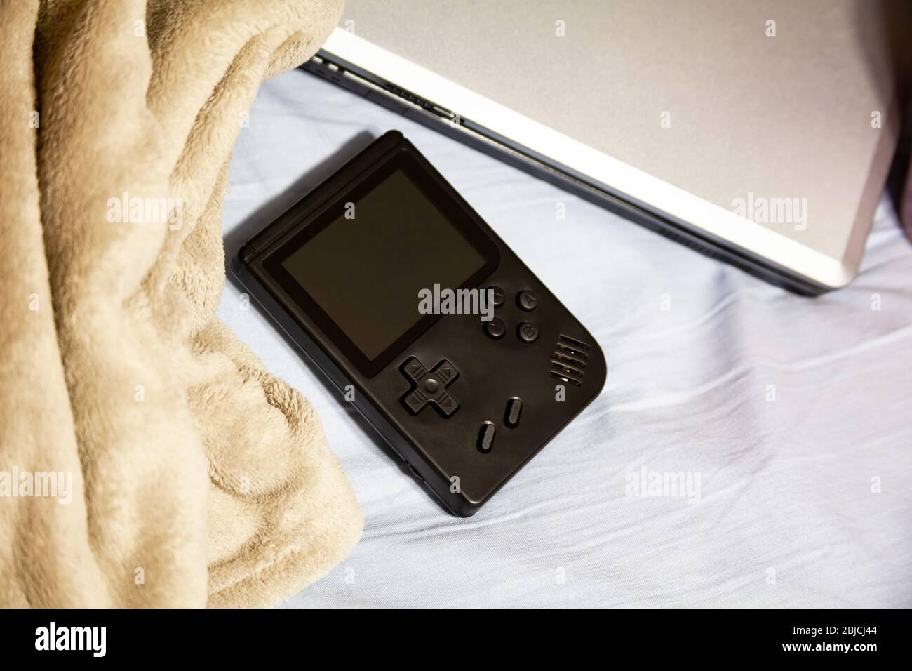 Console de jeux portable de poche, ordinateur portable et couverture confortable sur un lit non fabriqué avec des draps froissés. Activités de divertissement à domicile, ennui Banque D'Images