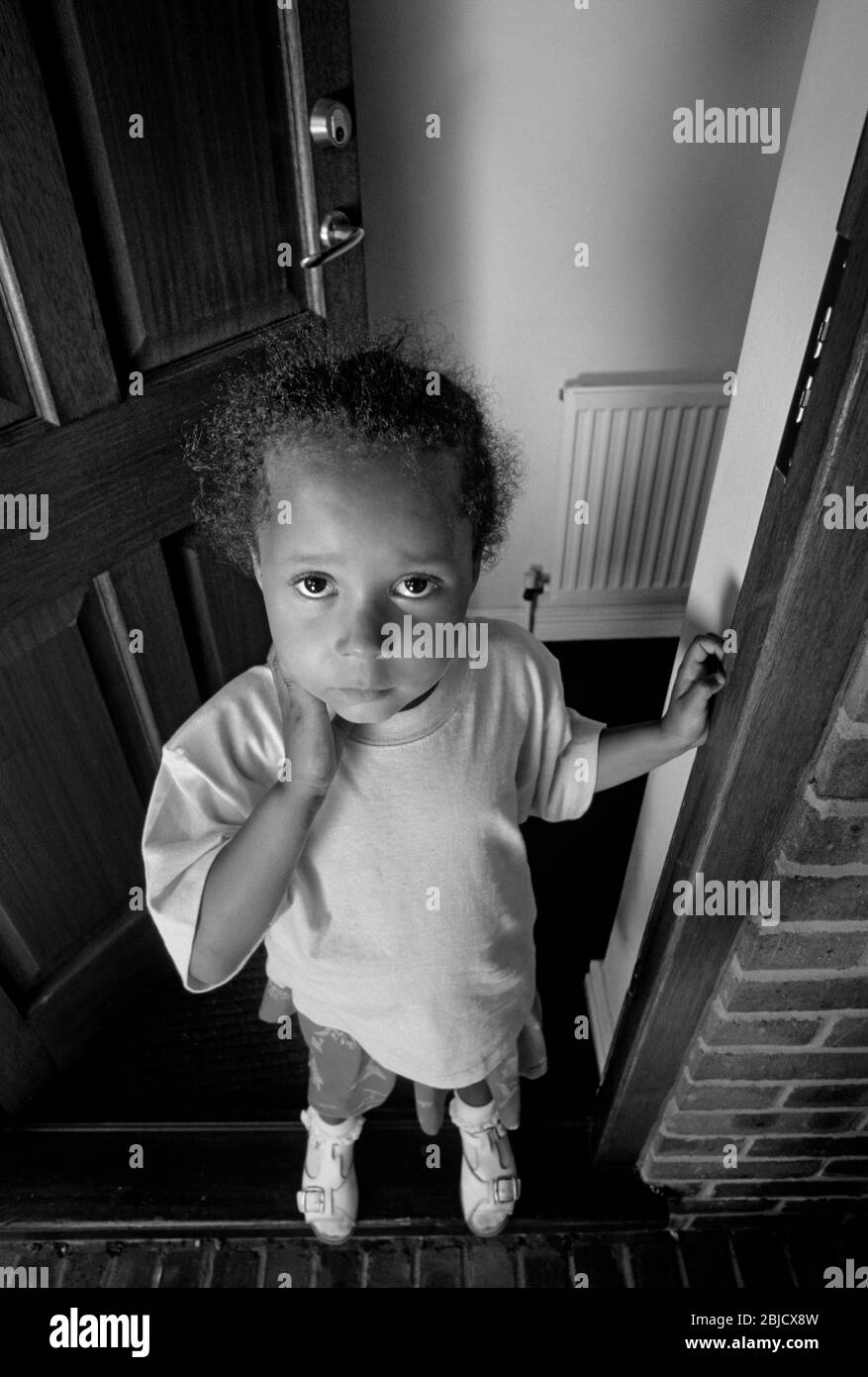 Bébé fille porte ouverte maison 3-5 ans mignon attrayant joli, se tient à l'entrée de porte ouverte de sa maison avec innocent triste pensive expression attrayante B & W. Banque D'Images