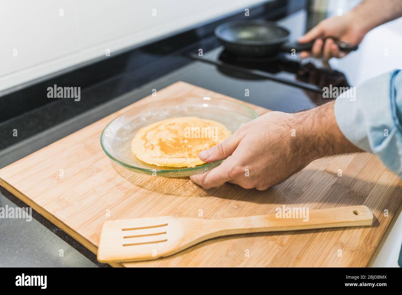 Homme cuisine dans la cuisine dans une chemise en denim. Un homme anonyme tient une poêle avec une crêpe sur une plaque Banque D'Images