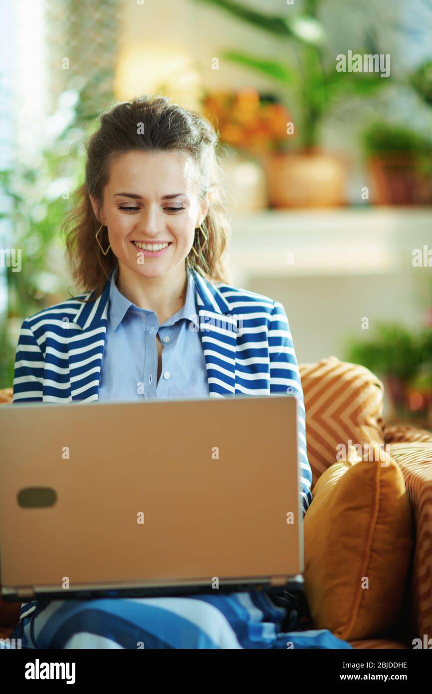 heureuse femme à la mode avec chemisier bleu et veste rayée assise sur canapé avec ordinateur portable dans la maison moderne en journée ensoleillée. Banque D'Images