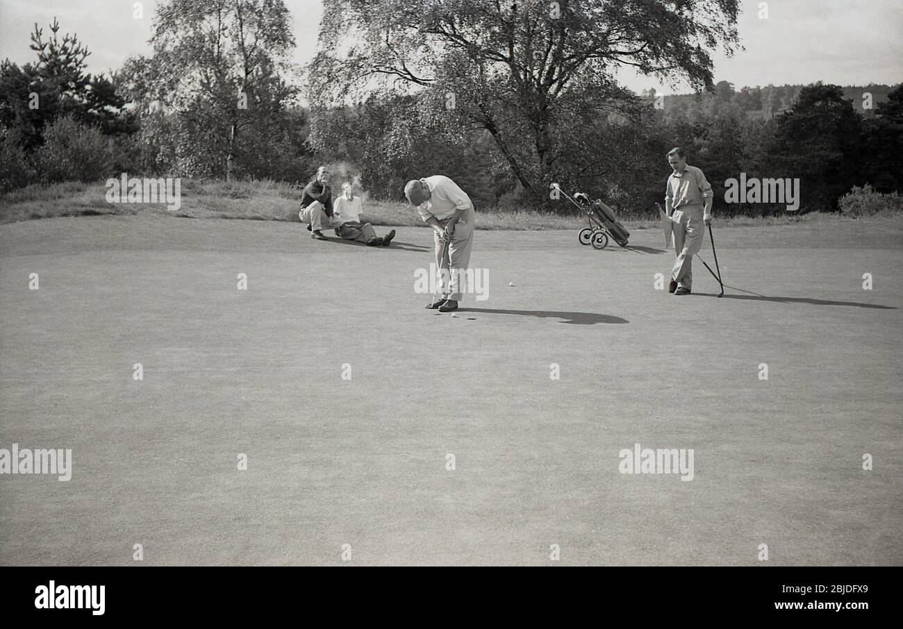 Années 60, historique, à l'extérieur sur un terrain de golf boisé, intérieur, deux golfeurs masculins sur un vert d'exercice, l'un tenant le drapeau, l'autre mettant, tandis que deux autres golfeurs dans le quatre-ball s'assoient par le vert ayant profité d'une cigarette, Angleterre, Royaume-Uni. On peut voir les modes, les clubs et un chariot de golf de l'époque. Banque D'Images