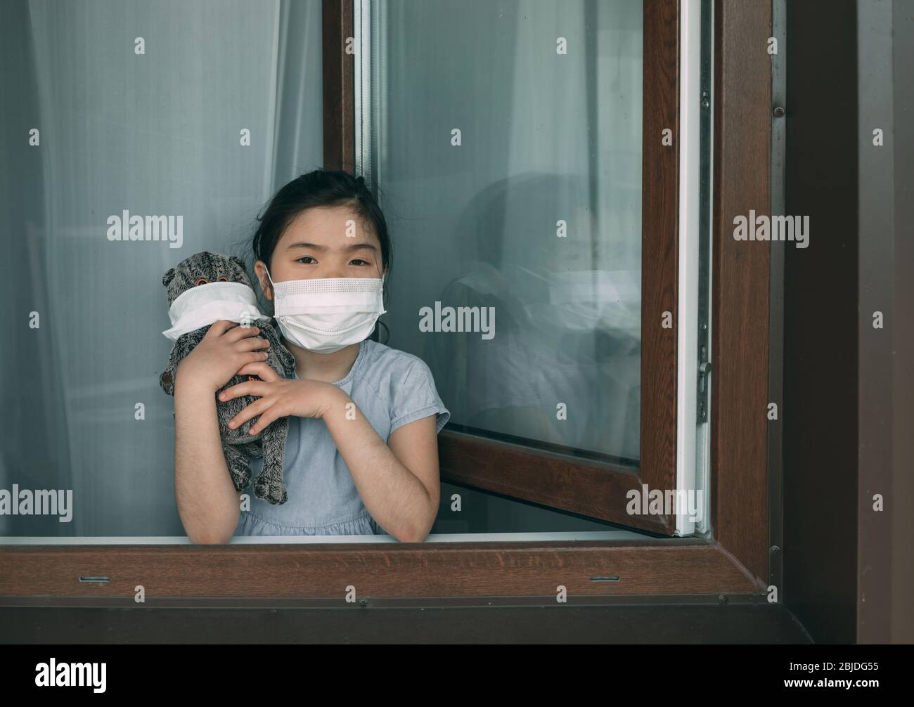 Une fille dans un masque avec un jouet farci se tient à la fenêtre ouverte de sa maison Banque D'Images