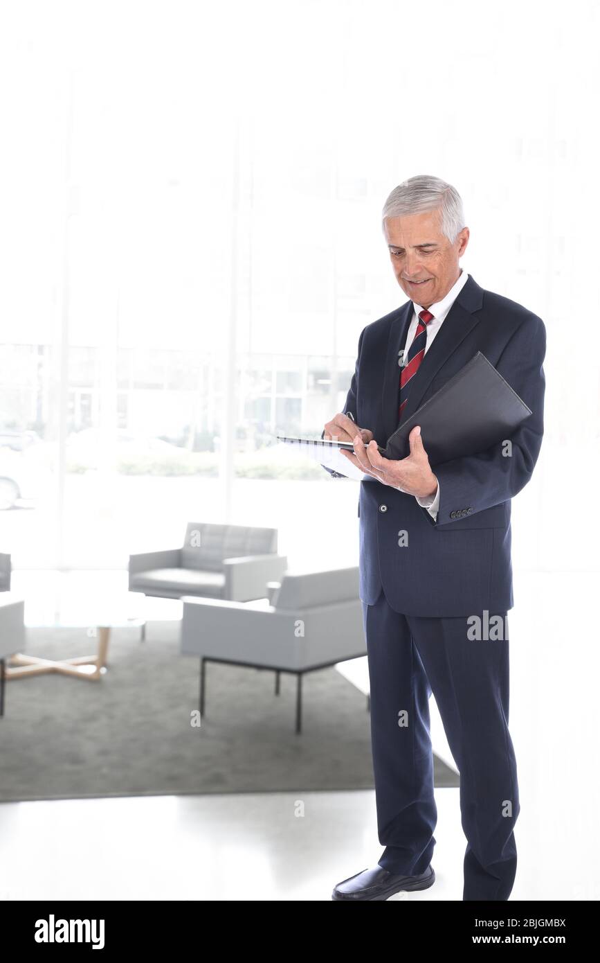 Homme d'affaires souriant et mûr debout dans un bâtiment moderne. L'homme écrit dans un dossier lié au cuir. Format vertical avec fond de touche Haut. Banque D'Images