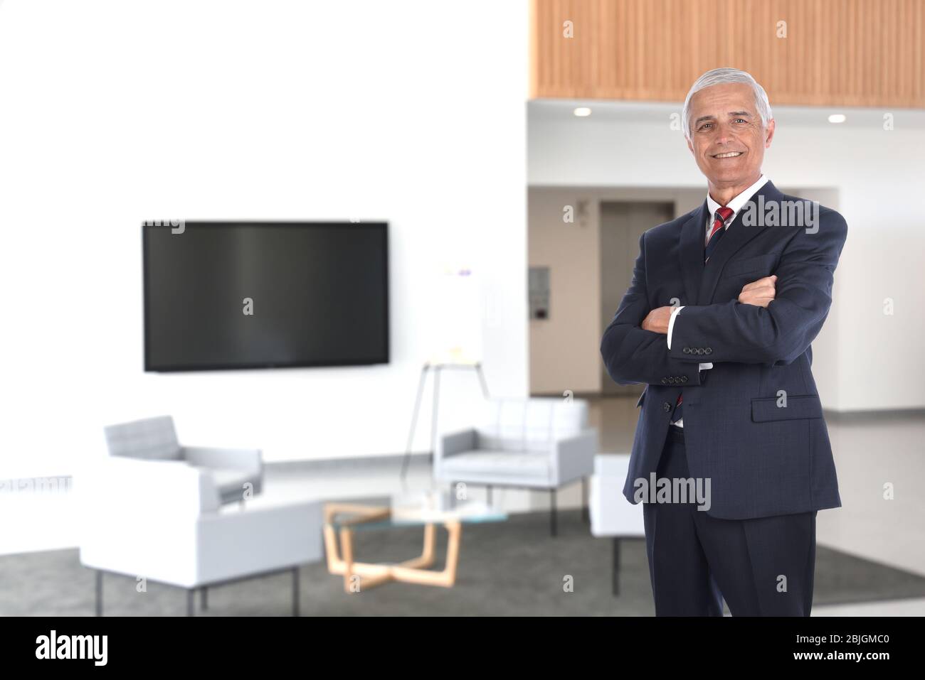 Homme d'affaires souriant et mûr, bras repliés dans un hall moderne de bureau avec télévision à écran plat. Format horizontal avec touche de fonction de touche haut Banque D'Images